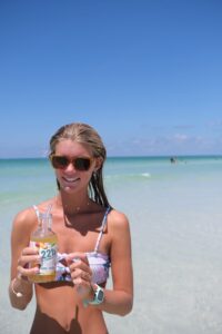 Chloe in summer bikini on beach with mango dandelion kombucha bottle